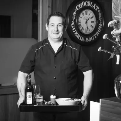 Le chef du restaurant de l'hôtel Stimotel souriant en uniforme de service tenant un plateau avec repas et boissons.