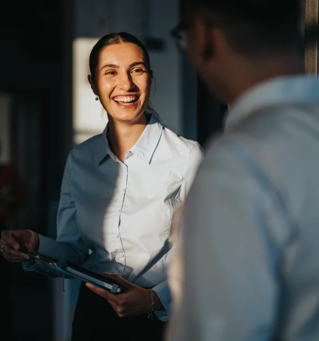 Femme souriante en chemise claire discutant avec un collègue dans un bureau faiblement éclairé.